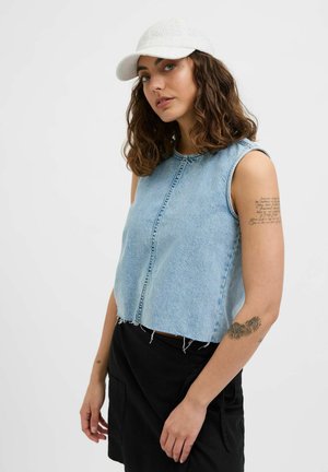 Kvinde med krøllet brunt hår, der bærer en hvid kasket, en ærmeløs lys blå denimtop, en sort nederdel og tatoveringer på sin venstre arm.