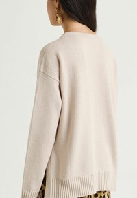 Beiger Strickpullover mit lockerem Schnitt, geripptem Saum und weiten Ärmeln. Die Textur ist glatt mit einem dezenten Glanz, seitliche Schlitze sind sichtbar.