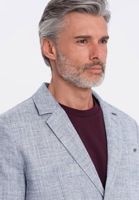 Hellgrauer Leinenblazer mit strukturiertem Stoff, Reverskragen und einer einzigen Brusttasche, getragen über einem dunkelrotbraunen Hemd.