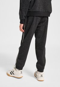 Schwarze, strukturierte Jogginghosen mit einem gesprenkelten Muster, versehen mit weißen Seitenstreifen. Dabei kombiniert mit weißen Sneakers mit schwarzen Akzenten und einem Logo.