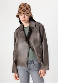 Brun imitert lær bikerjakke med glidelås lommer, båret over en grønn og hvitstripet skjorte. Svart mønstret bøttehatt. Grå jeans.