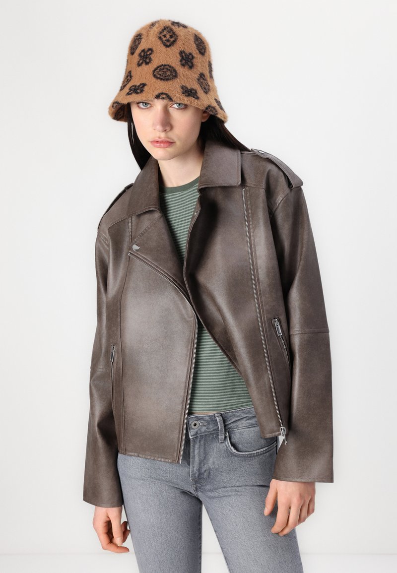 Giacca biker in finta pelle marrone con tasche zippate, indossata sopra una camicia a righe verde e bianca. Cappello a secchiello nero con fantasia. Jeans grigi.