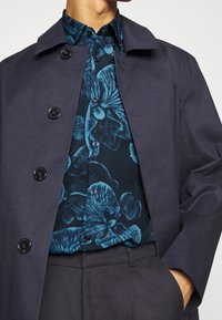 Marine blazer met donkere knopen boven een blauw met bloemenpatroon overhemd. Het overhemd heeft grote orchidee-ontwerpen. Grijze broek maakt de outfit compleet.