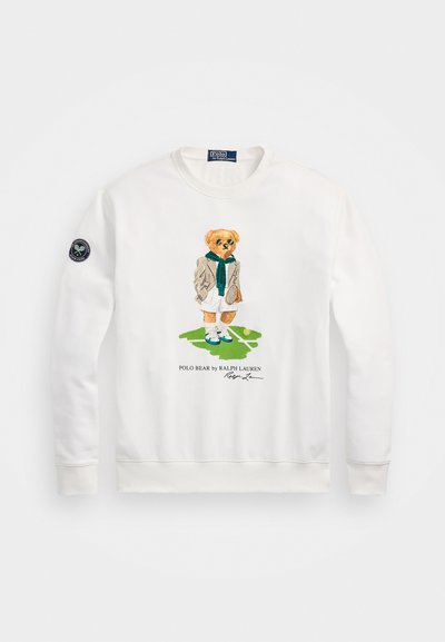 Sweatshirt branco com um urso de cartoon a usar uma jaqueta e um cachecol, segurando uma raquete de ténis. Inclui um logo em forma de patch na manga.