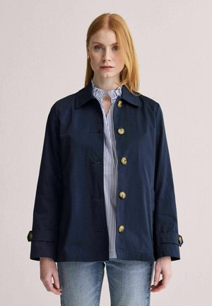 Rødhåret kvinde iført en marineblå knapjakke over en blå og hvidstribet skjorte og lyseblå jeans, stående foran en ensfarvet baggrund.