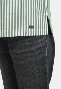 Camicia a righe verdi e bianche con un'etichetta, abbinata a jeans grigio scuro. La camicia presenta un orlo dritto con spacchi laterali.