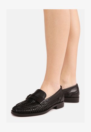 Melni pinuma raksta ādas loafers kurpes valkātas uz kailām kājām, ar zemu papēdi un noapaļotu purngalu, attēlotas uz balta fona.