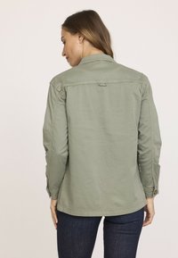 Veste chemise vert olive avec un col rabattable, des poignets boutonnés et un ourlet arrondi. Fabriquée en tissu léger avec une texture lisse.