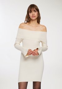 Abito a spalle scoperte in maglia di colore crema, fitted con maniche a campana e orlo che arriva al ginocchio. Texture morbida e design senza cuciture.