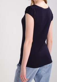 Mujer de piel clara con una camiseta negra ajustada de manga corta y jeans de mezclilla azul claros, mostrada de espaldas contra un fondo simple.