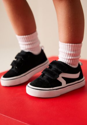 Enfant portant des baskets noires et blanches avec des bandes velcro et des chaussettes blanches côtelées, debout sur une surface rouge.