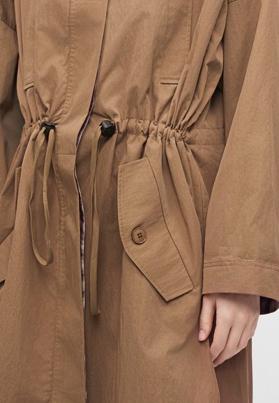 Manteau marron avec cordons resserrés à la taille, poche à rabat boutonnée et main partiellement visible sur le côté.