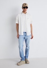 Camicia bianca con bottoni sopra una t-shirt bianca, jeans azzurri chiari, berretto beige, occhiali da sole scuri e sneakers marrone chiaro. Texture lisce.