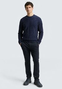 Navy blue knitted sweater with speckled detailing, paired with dark trousers and black sneakers. Model stands with hands in pockets.
