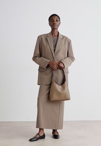 Blazer beige sartoriale con decorazioni sulle spalle, abbinato a una gonna fitted coordinata. Il modello tiene una borsa hobo marrone, indossando ballerine nere.