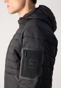 Sānu skats uz personu, kas valkā melnu pieņemamu jaku ar rāvējslēdzēja kabatu un izspiestu BOSS logo uz augšējā piedurknes.