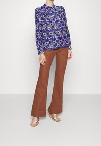 Blouse à manches longues à fleurs bleu et blanc associée à un pantalon évasé marron. Elle présente un col à ouverture clé et des sandales à talons texturées.