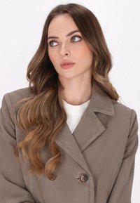 Khaki dwurzędowy trencz z kołnierzem z wycięciem, ozdobiony brązowymi guzikami i gładką teksturą materiału, noszony na białej bluzce.