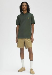 Polo shirt verde scuro con logo dorato, abbinato a pantaloni corti beige. Tessuto testurizzato, maniche corte e calzini bianchi con dettagli neri.