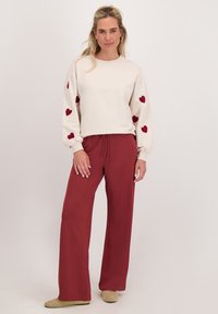 Crème sweatshirt met rode hartafdrukken op de mouwen, gecombineerd met bordeaux brede broek, gepresenteerd op een persoon tegen een witte achtergrond.