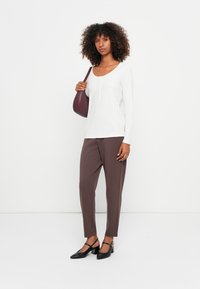 Vrouw met krullend haar, gekleed in een witte lange mouwen top, bruine broek, zwarte hakken en een bordeauxrode schoudertas, staande tegen een witte achtergrond.