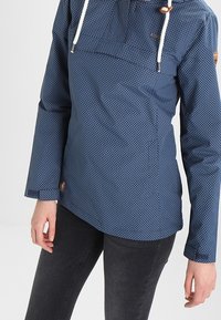 Granatowy pulower w białe kropki, z pół-zamkiem przy szyi, sznurkami do regulacji i bocznymi rozporkami. Posiada znak firmowy na dolnej lewej stronie.