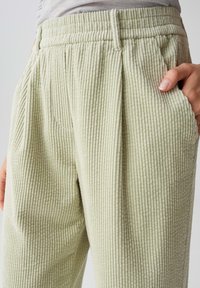 Pantaloni in corduroy verde chiaro con texture a righe verticali, dotati di vita e tasche laterali. Vestibilità ampia con pieghe sul davanti.