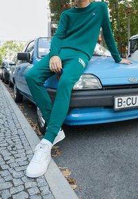Zielony sweter i dopasowane dresy z miękkiego materiału, z logo. Białe sneakersy o teksturowanej powierzchni, leżące na masce niebieskiego samochodu.
