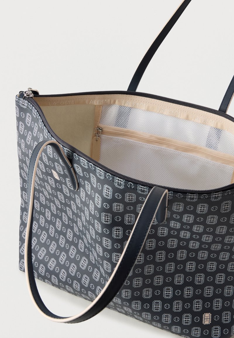Marineblaue Tragetasche mit weißem geometrischem Muster, schwarzen Griffen, beigem Innenfutter und innerer Reißverschluss-Netztasche.