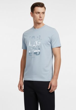 Junger Mann, der ein hellblaues Kurzarm-T-Shirt mit reflektierendem "KARL LAGERFELD" Schriftzug und schwarze Hose trägt, blickt nach links.