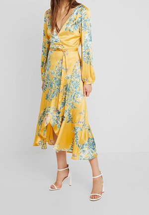 Femme portant une robe portefeuille jaune à fleurs avec manches longues et ourlet volanté, assortie à des sandales blanches à talons hauts sur un fond uni.