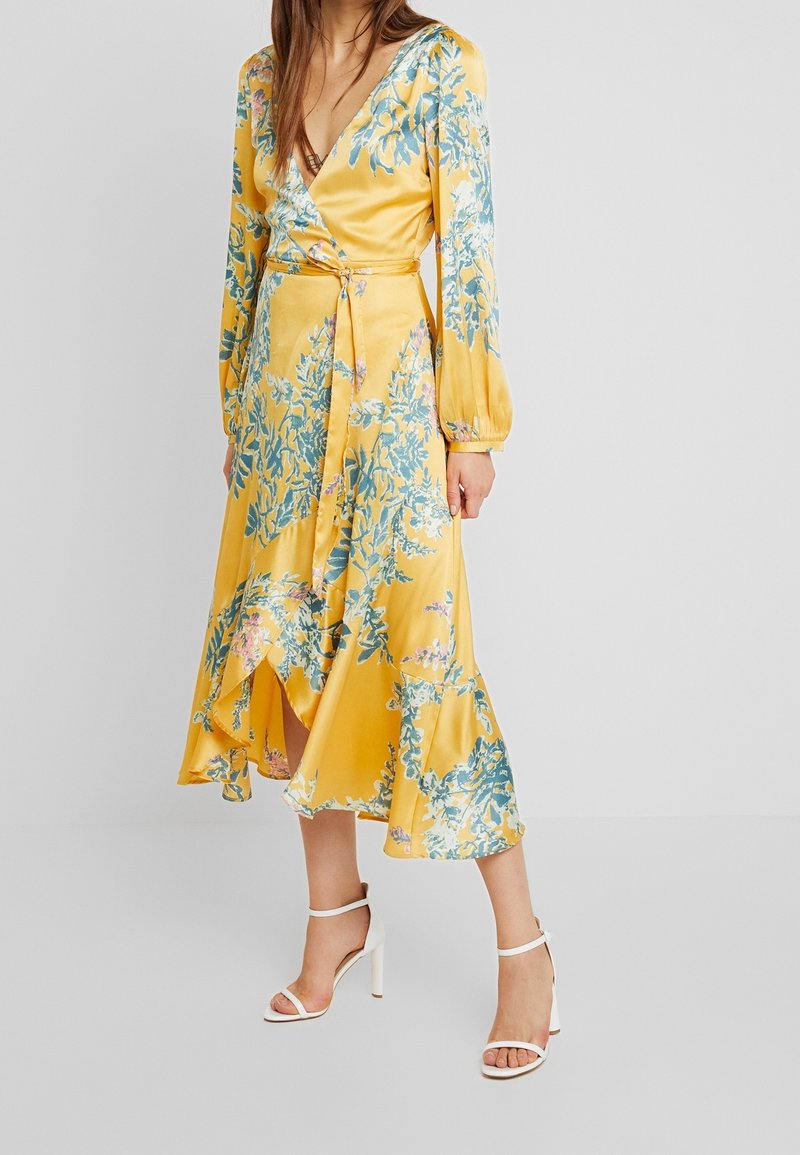 Femme portant une robe portefeuille jaune à fleurs avec manches longues et ourlet volanté, assortie à des sandales blanches à talons hauts sur un fond uni.
