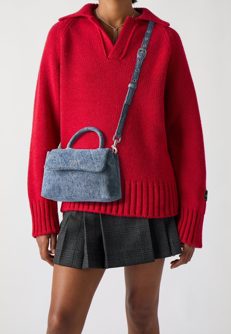 Personne portant un pull en maille rouge et une jupe plissée à carreaux noirs, portant un petit sac à main en jean bleu avec une poignée supérieure et une bandoulière réglable.