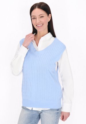 Žena se usmívá, má na sobě světle modrý pletený svetřík bez rukávů přes bílou košili s knoflíky a světle modré džíny, stojí před bílým pozadím.