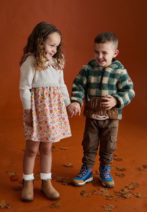 Zwei Kinder halten Händchen vor einem orangefarbenen Hintergrund. Das Mädchen trägt ein Blumenkleid und einen beigefarbenen Strickpullover; der Junge hat einen grün-karierte Fleecejacke und braune Hosen an.