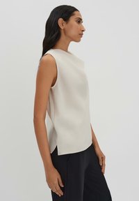 Sleeveless cream top with a high neckline, soft texture, and curved sides, paired with loose black pants. Minimalistic design.