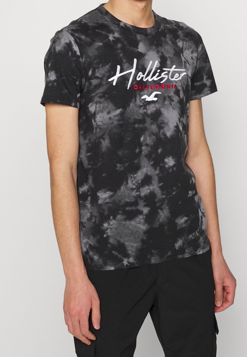 Svart tie-dye t-shirt med korta ärmar, prydd med en vit skriptlogotyp "Hollister" och "California" i röd färg. Tillverkas av bomull, avslappnad passform.