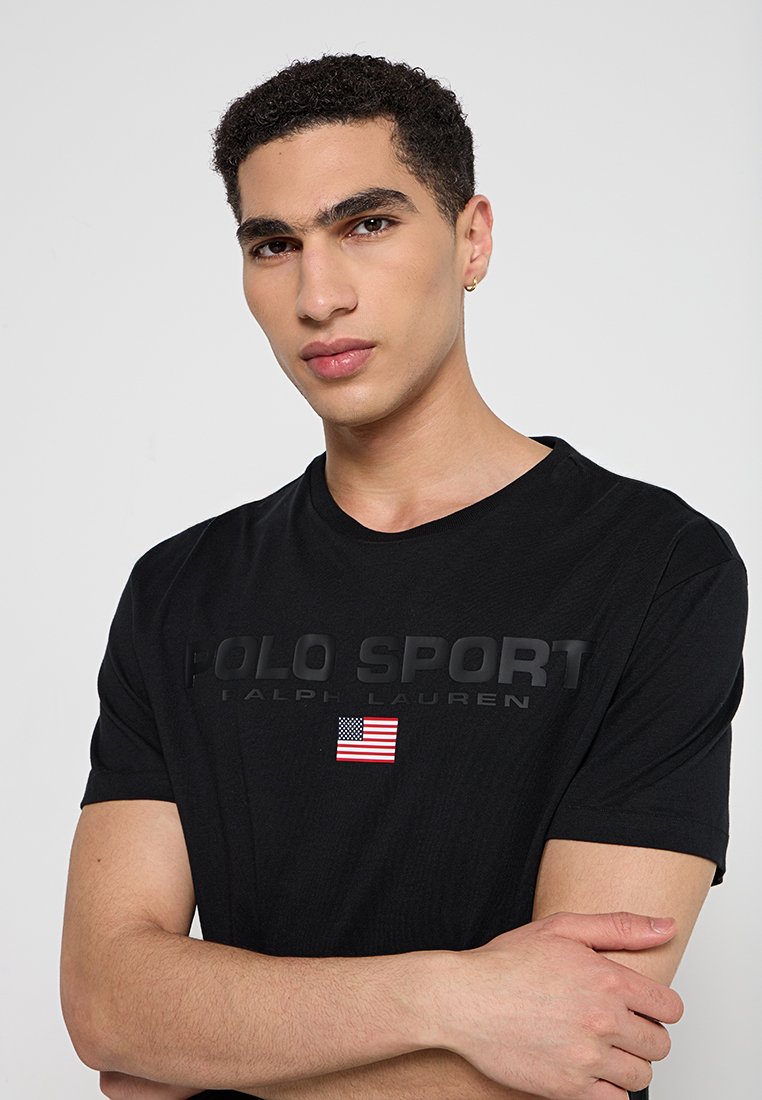 T-shirt nera in cotone con la scritta "POLO SPORT" in rilievo, completa di un piccolo emblema della bandiera americana. Maniche corte e design a girocollo.