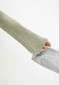 Manica di un maglione verde chiaro, con texture e un ampio polsino, che tiene un sacchetto di rete argentata con una superficie liscia.