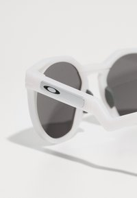 Gafas de sol con montura octagonal blanca, lentes oscuros, logo plateado en la patilla y acentos de goma para mejorar el agarre. Textura brillante en toda la superficie.