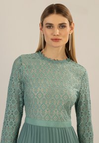 Un vestito a maniche lunghe di color azzurro chiaro presenta un motivo in pizzo con diamanti testurizzati, un collo a cappuccio e una gonna plissettata.