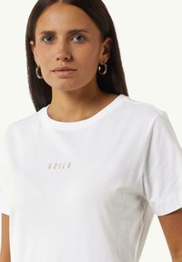 White cotton t-shirt featuring short sleeves and a crew neck, adorned with “VOILÀ” in light beige embroidery at the chest.