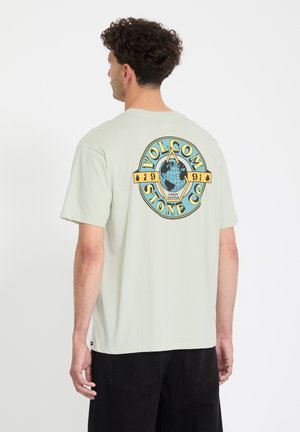 Man met krullend haar, gekleed in een lichtgrijs t-shirt met een kleurrijk Volcom Stone Co. 1991 globe-logo op de achterkant, gecombineerd met zwarte shorts.