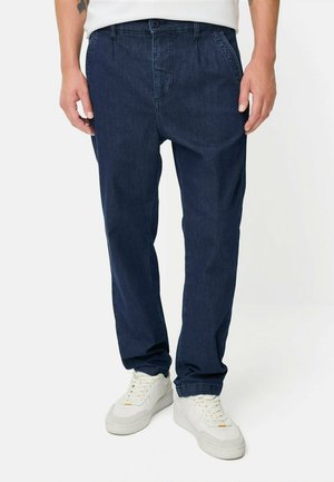 Mand iført mørkeblå denimbukser og hvide sneakers med en hvid skjorte, stående mod en ensfarvet hvid baggrund.