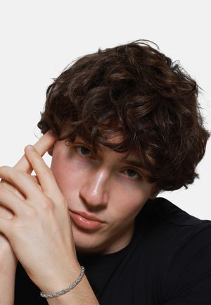 Personne avec des cheveux bruns bouclés portant une chemise noire, les mains tenues près du visage. Un bracelet en argent tressé est porté à un poignet. Fond blanc.