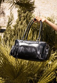Black leather handbag with dual top handles, multiple zippered pockets, smooth texture, rectangular shape, and glossy finish.
