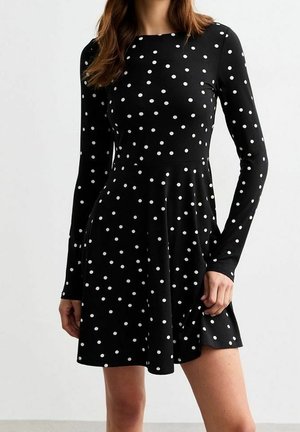 Kobieta w czarnej sukience z długim rękawem w białe kropki, delikatnie trzymająca spódnicę jedną ręką.