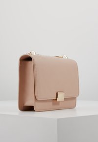 Borsa a mano in pelle rosa chiaro con dettagli in oro, chiusura quadrata, forma strutturata e tracolla a catena, posata su un piedistallo grigio chiaro.