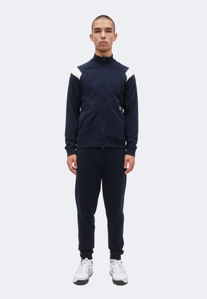 Tânăr stă cu fața înainte purtând o geacă albastru închis cu fermoar, cu accente albe pe umeri, pantaloni asortați și adidași albi.