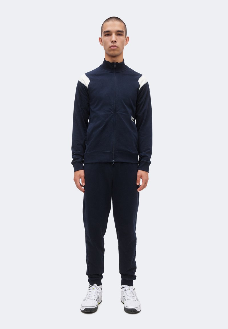 Νέος άνδρας στέκεται προς τα εμπρός φορώντας μπλε σκούρο μπουφάν με φερμουάρ και λευκές λεπτομέρειες στους ώμους, ταιριαστό παντελόνι και λευκά αθλητικά παπούτσια.